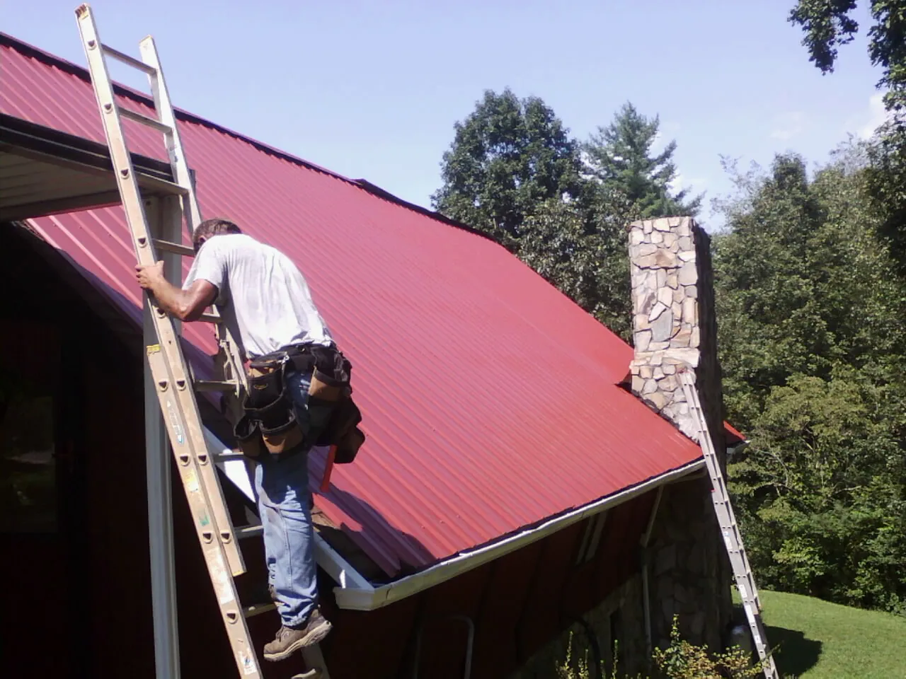 Roofer on a ladder beside a red metal roof and stone chimney — Soffit & Fascia Repair by Roofing Brothers in Mount Laurel